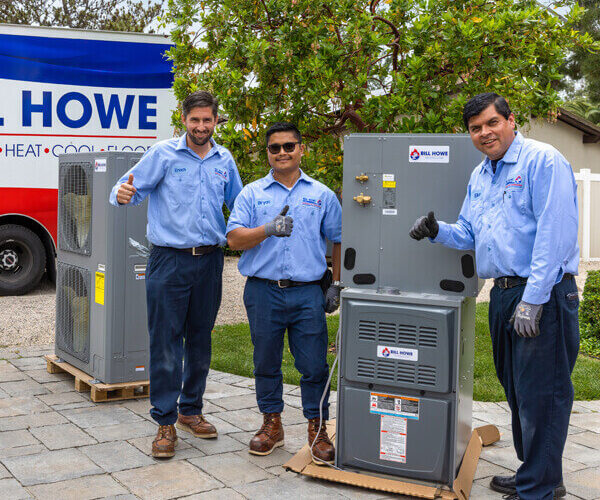 Three smiling HVAC technicians wearing Bill Howe uniforms standing near HVAC equipment with their thumbs up.